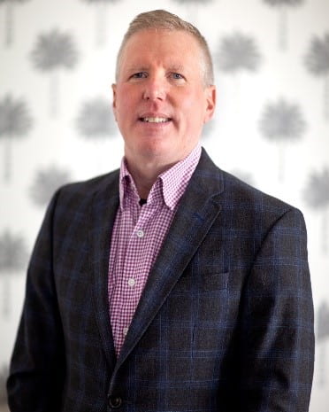 A professional photo of Chris Cowen, a caucasian man with blond greying hair and blue eyes is dressed in a black blazer with blue plaid paired with a white and red checked button up shirt.  The background is white with faint grey palm trees.